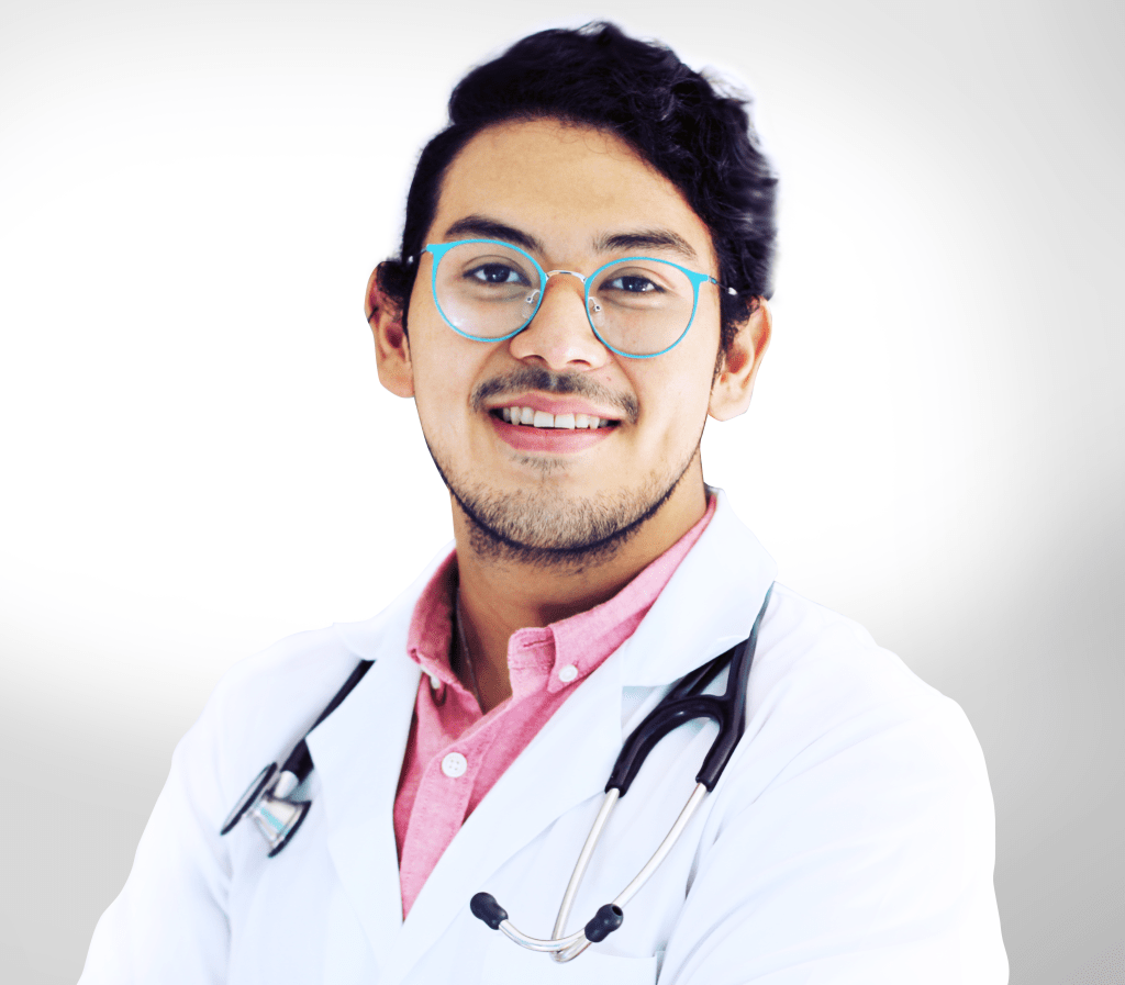 Juan wearing a white lab coat with a stethoscope hanging from his shoulders. He is staring at the camera with a smile. Juan is wearing light blue glasses and his hair is tied in the back.
